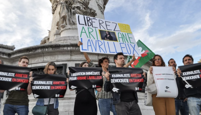 A group of people stand in front of a monument holding banners in support of journalist Ariane Lavrilleux. The signs read messages such as “Journaliste arrêtée, démocratie en danger” (“Journalist arrested, democracy in danger”), “Libération immédiate pour Ariane Lavrilleux” (“Immediate release for Ariane Lavrilleux”), and “Secret des sources, défense d’y toucher” (“Protection of sources, do not interfere”). One person holds a large sign with a photo of Lavrilleux and the words “Libérez Ariane Lavrilleux.”
